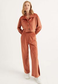 Rostfarbene Cordjacke mit Cropped-Schnitt und Knöpfen sowie passenden elastischen Hosen mit seitlichen Schlitzen. Das Model trägt weiße Sneaker.