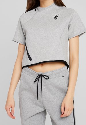 Femme portant un t-shirt Nike court gris clair et un jogging assorti avec des cordons noirs, debout devant un fond uni.