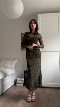 Robe maxi transparente vert olive avec motifs floraux, manches longues et silhouette ajustée. Accessoirisée avec des sandales noires à lanières et un sac.