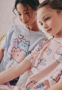 Två tjejer modellerar pastellfärgade t-shirts: en blå med tecknade katter och hjärtan, den andra rosa med färgglada katter och stjärnor. Mjuk tyg, avslappnad passform.