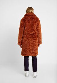 Lang, fluffy frakke i brændt orange med bred krave og hele ærmer. Båret over stribede bukser og hvide sneakers.