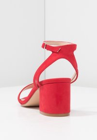 Sandale à talon en daim rouge avec un talon bloc et une lanière à la cheville dotée d'une boucle en argent. Design minimaliste avec des textures lisses et des lignes épurées.