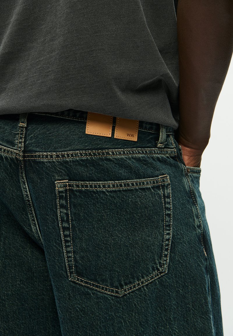 Dark-skinned person wearing dark blue denim jeans and a dark grey shirt, with hand tucked into back pocket on a white background.