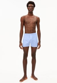 Helle blaue Shorts aus weichem Stoff, mit einem elastischen Bund und lockerer Passform, getragen von einem männlichen Modell mit baren Füßen.
