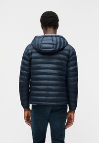 Άνδρας που φοράει σκούρο μπλε μπουφάν με κουκούλα και σκούρο τζιν, στέκεται με την πλάτη προς τα έξω σε απλό φωτεινό φόντο.