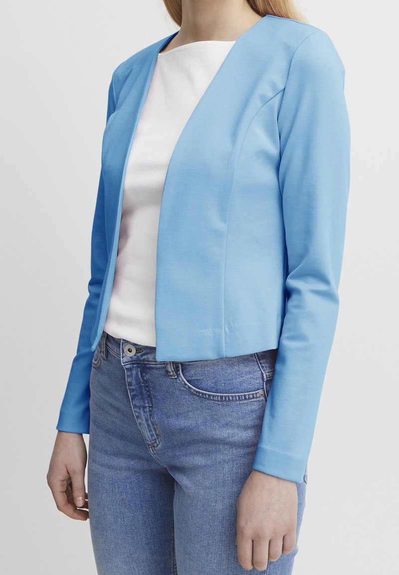 Femme portant un blazer bleu clair ajusté sur un haut blanc et un jean bleu, debout avec les bras détendus le long du corps.