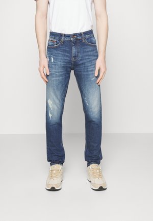 Uomo con jeans blu sbiaditi e consumati, t-shirt bianca e scarpe da ginnastica beige, in piedi contro uno sfondo chiaro e semplice.