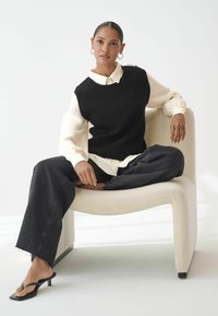 Un pull sans manches noir en tricot sur une blouse crème, associé à un jean noir à jambes larges et des sandales à talons noires. Assise sur une chaise de couleur claire.