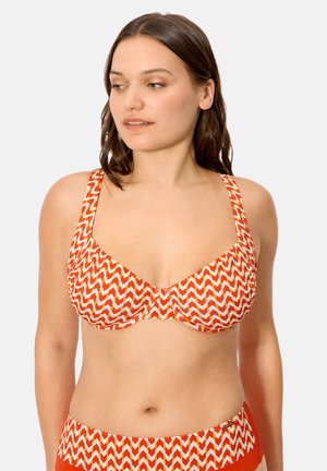 Mujer con bikini en patrón zigzag naranja y blanco, mirando ligeramente hacia la izquierda contra un fondo claro y sencillo.