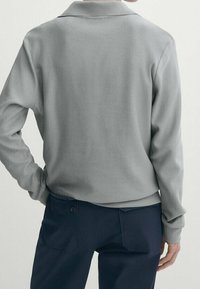 Szara koszula z długim rękawem o fakturowanej tkaninie, widoczna od tyłu, w połączeniu z granatowymi spodniami wyposażonymi w boczne kieszenie.