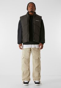 Zwarte gewatteerde vest over een zwart sweatshirt, gecombineerd met beige cargobroeken met meerdere zakken. Schoenen zijn zwart met witte accenten.