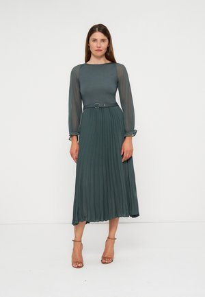 Femme portant une longue robe plissée vert foncé avec des manches longues transparentes et une ceinture, debout sur un fond blanc uni.