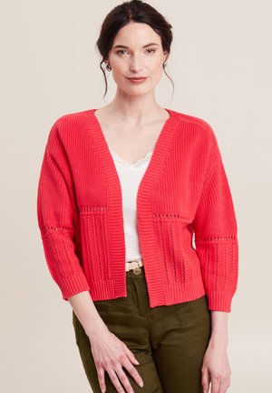 Koraalkleurige gebreide cardigan met een V-hals, driekwartmouwen en open geweven details, in combinatie met een witte tanktop en groene broek.