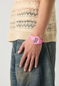 Rosa digital klokke med silikonrem og rund urskive, med hvit ramme og klar skjerm, båret på en hånd mot denim.