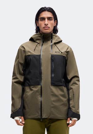 Homme aux cheveux foncés mi-longs porte une veste à capuche vert olive et noire avec une fermeture éclair frontale et une poche poitrine zippée sur un fond clair.