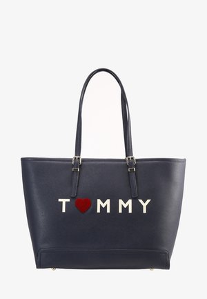 Granatowa torba na ramię wykonana z materiału syntetycznego, z białym napisem „TOMMY” i czerwonym akcentem w kształcie serca. Podwójne uchwyty i usztywniona konstrukcja.