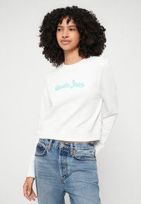 Λευκή κοντή μπλούζα με την τουρκουάζ λογότυπο "Guess Jeans", στρογγυλή λαιμόκοψη, ριμπ μανσέτες και στρίφωμα; συνδυασμένη με ανοιχτό μπλε τζιν παντελόνι.