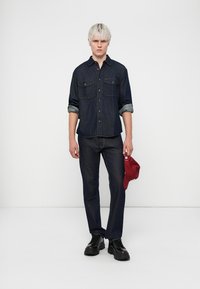 Mørk denimskjorte med knappestolpe og to brystlommer, parret med mørke denimbukser. Modell holder en rød caps og har på seg svarte sko.