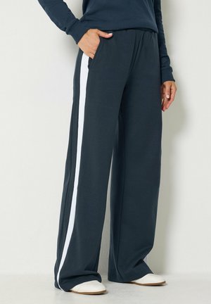 Personne portant un pantalon large foncé avec des bandes blanches sur les côtés et des chaussures blanches, main reposant dans la poche du pantalon.
