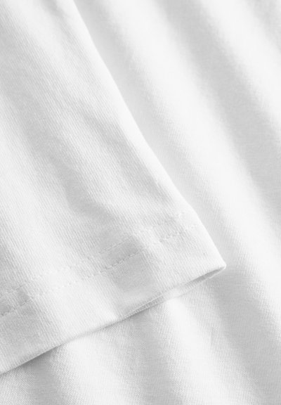 Tessuto di cotone bianco con una texture liscia, che mostra un bordo piegato e un dettaglio di cucitura lungo l'orlo. Senza motivi o accenti presenti.