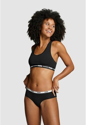 Smiling woman with curly hair wearing black Puma sports bra and matching briefs, adjusting waistband, standing against plain light background.