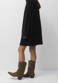 Des bottes en suede vert olive à hauteur de genoux avec un bout pointu, un talon légèrement évasé et des détails de couture sur le côté, associées à une robe noire fluide.