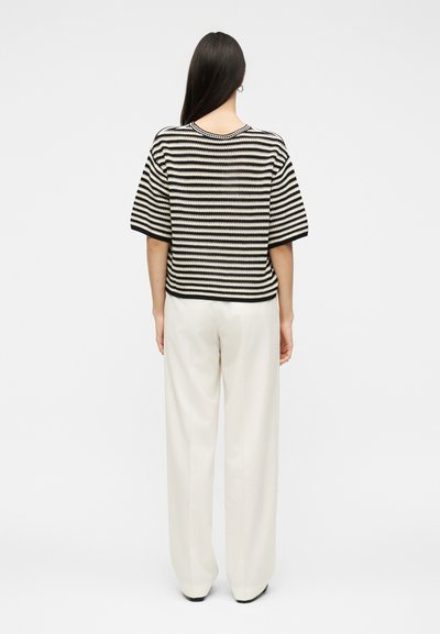 Γυναίκα με μακριά σκούρα μαλλιά που φοράει ασπρόμαυρο ριγέ κοντομάνικο πουλόβερ και άνετο λευκό παντελόνι, στέκεται με την πλάτη της προς το απλό φόντο.