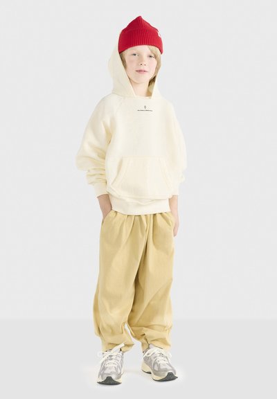 Felpa crema con tasca frontale, logo sul petto; pantaloni beige larghi; berretto di maglia rosso; sneakers grigio e bianche con dettagli neri.