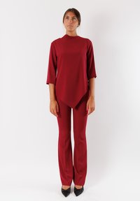Top rosso con colletto alla cinese, maniche a tre quarti e vestibilità ampia, abbinato a pantaloni a zampa coordinati e tacchi neri.