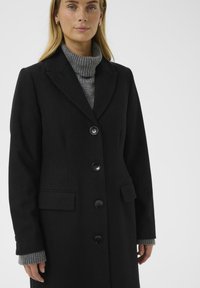 Woman wearing a black buttoned coat over a gray turtleneck sweater, with earrings visible, standing against a white background.