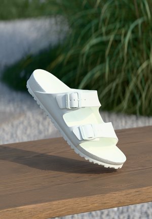 Biała sandałka na wsuwkę z dwoma regulowanymi paskami z klamrami, spoczywająca na drewnianej powierzchni na zewnątrz, w tle zielone rośliny.