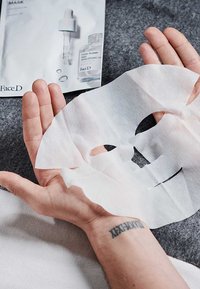 Maschera viso in tessuto bianco tenuta tra le mani, con aperture per occhi e bocca ritagliate. Lo sfondo presenta il packaging del prodotto con contagocce e siero.
