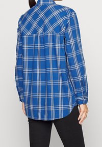 Camisa de cuadros azul con mangas largas, que presenta Rayas blancas y azul oscuro, un dobladillo redondeado y un diseño de espalda plisada.