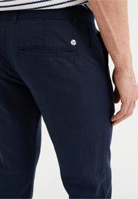 Mörkblå byxor med bakficka och silverfärgad märkesknapp, bärs av person i randig skjorta, hand synlig vid sidan.