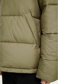 Olivgrön vadderad jacka med en strukturerad yta, oversize-silhuett och sidoficka med en diagonal söm. Detaljer inkluderar en foderkant vid ärmslutet.