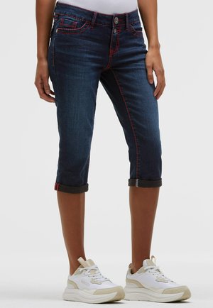 Woman wearing dark blue cropped jeans with red stitching and white sneakers with beige accents, standing against a plain background.