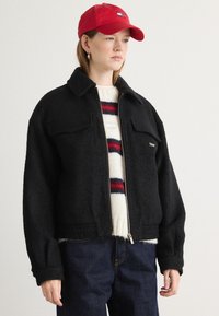 Sort fleecejakke med lynlås og brystlommer, båret over en cremefarvet sweater med røde og blå striber, parret med mørke jeans og en rød kasket.