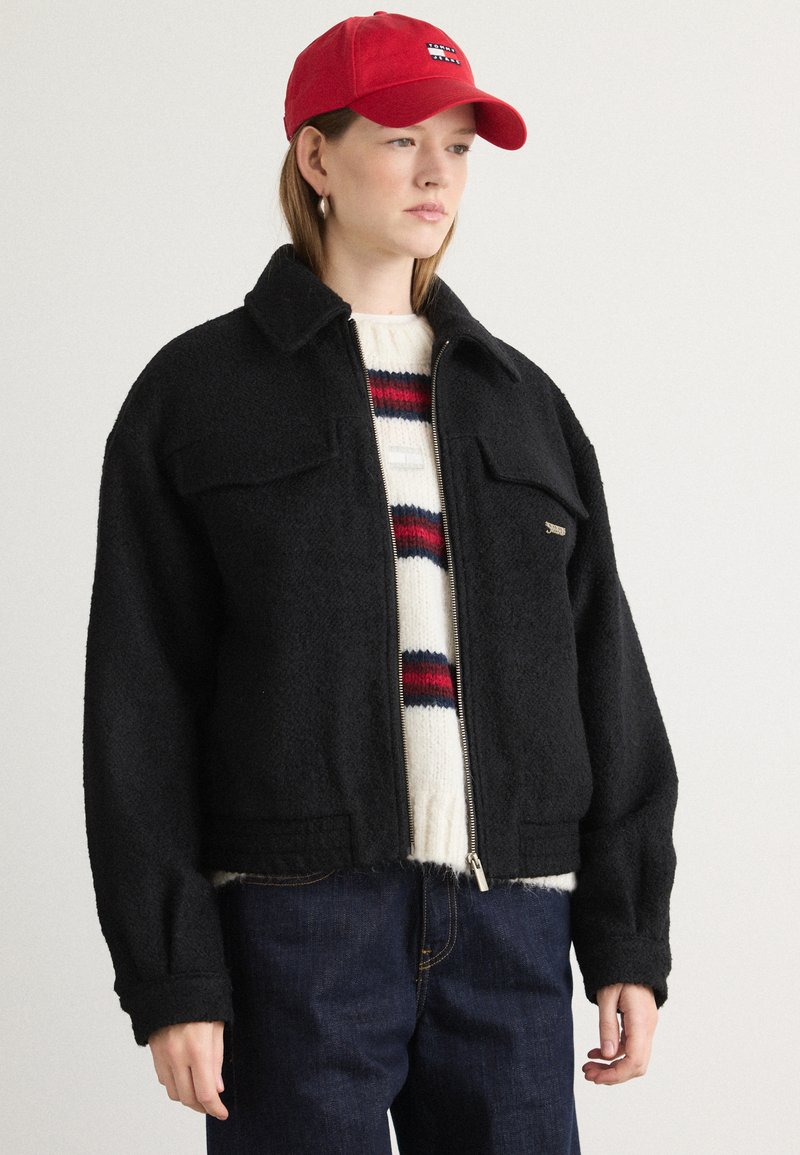 Sort fleecejakke med lynlås og brystlommer, båret over en cremefarvet sweater med røde og blå striber, parret med mørke jeans og en rød kasket.