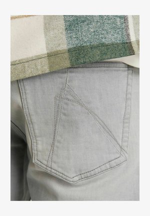 Pantalones de mezclilla grises con un diseño único de bolsillos traseros en ángulo, combinados con una camisa de franela a cuadros en tonos beige y verde.