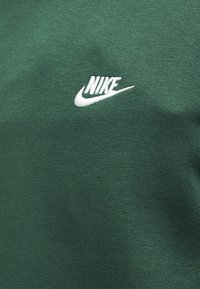 Λευκό λογότυπο της Nike κεντημένο σε σκουροπράσινο υφασμάτινο ύφασμα με υφή, πιθανώς φούτερ ή μπλουζάκι.