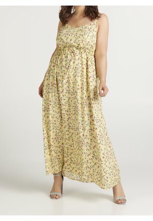 Femme portant une robe longue jaune à fleurs avec fines bretelles et ceinture à la taille, associée à des sandales à talons hauts gris clair.