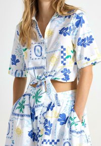 Kvinna iklädd en vit tvådelad klädnad med blå, gröna och gula tropiska och geometriska mönster, med en knytcrop-top och en högmidjad kjol.