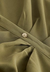 Olivenfarbenes, strukturiertes Gewebe mit Falten und einem dekorativen goldenen Knopf, der die Mitte betont. Glattes Material, minimalistisches Design.
