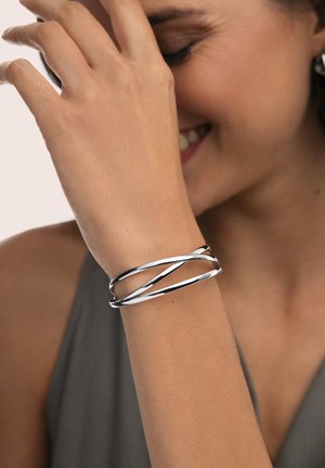 Mujer sonriente con los ojos cerrados que lleva una pulsera de plata torcida en la muñeca, con la mano cubriéndose parcialmente el rostro.