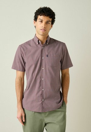 Young man wearing short-sleeve maroon and white checkered shirt with button-down collar and light green pants, standing against plain background.