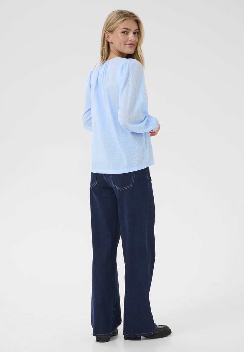 Hellblaue Bluse mit strukturiertem Punktmuster, leicht gepufften Ärmeln, kombiniert mit dunkelblauen weit geschnittenen Jeans und schwarzen Schuhen.