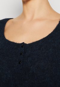 Námořnicky modrý svetr z vlněné směsi se širokým výstřihem, který má čtyři černé knoflíky a strukturovaný povrch. Detailní záběr, který zdůrazňuje texturu látky.