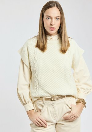 Une femme aux cheveux bruns lisses porte un pull sans manches en tricot torsadé crème sur une blouse à manches longues, un pantalon léger, une ceinture et des bracelets.