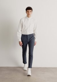 Camicia bianca a maniche lunghe con taschino, abbinata a pantaloni slim-fit grigio scuro e sneakers bianche, indossata da un modello su uno sfondo neutro.