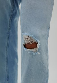 Vaqueros de mezclilla azul claro con un agujero desgastado en la rodilla, mostrando bordes deshilachados. Textura suave, diseño ajustado con detalles de costura estándar.